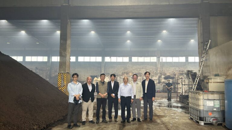 A moment of recognition: the core project team and visiting collaborators stand together inside the biochar facility. Behind them, bags of processed material and raw input show the scale of operations — a living example of innovation built from the ground up.