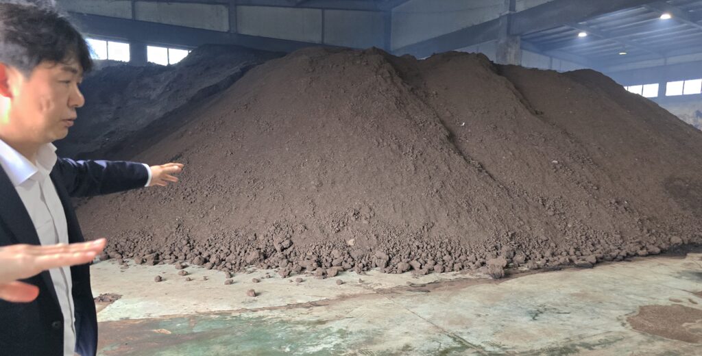 At the heart of the operation: a large pile of raw manure material inside the processing facility — the very starting point of the transformation into high-quality granular biochar. A representative gestures toward the scale and consistency of the input feedstock, carefully managed before entering fermentation and pyrolysis stages.